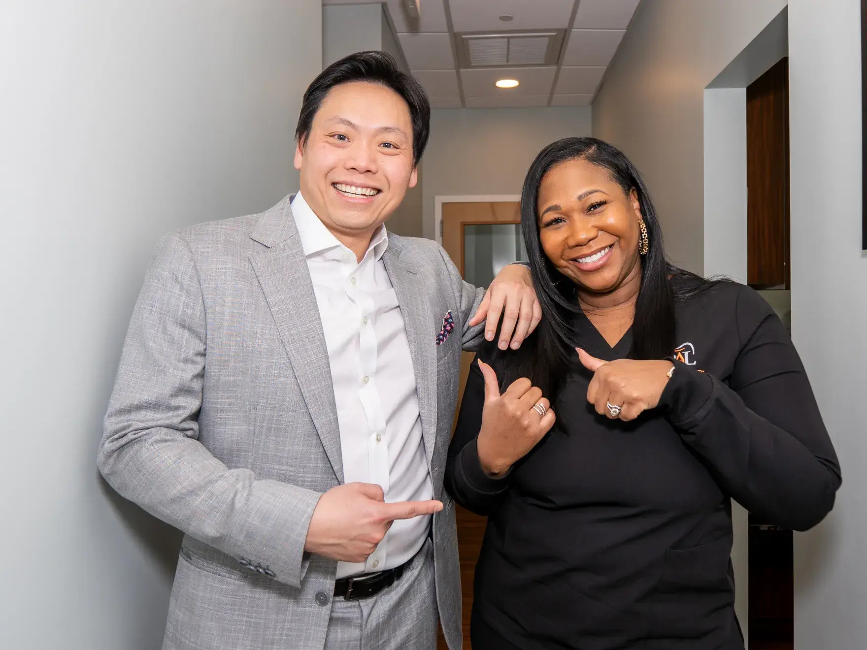 A man and a woman smile, giving thumbs up, standing side by side in a hallway.