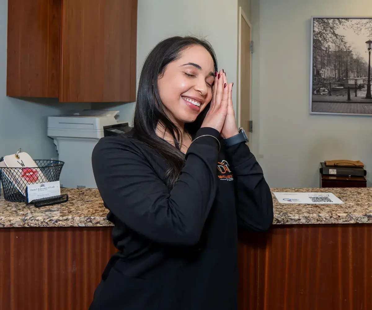 A woman in a black sweater stands at a reception desk, smiling with her hands together.