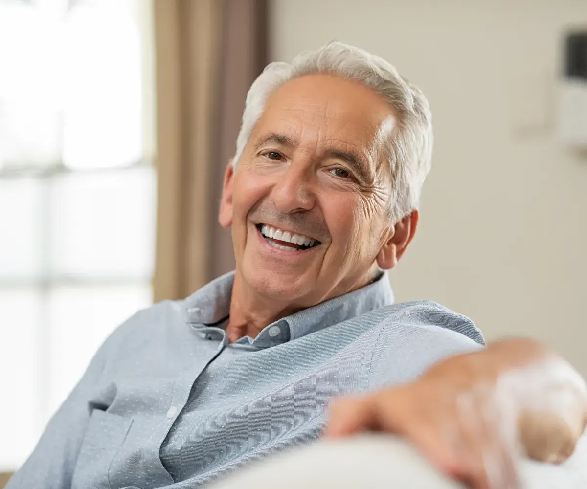 Smiling older man in a light blue shirt sitting indoors.
