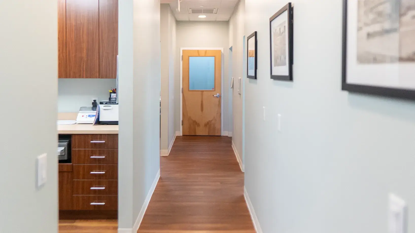 A hallway with light walls, wooden floor, closed wooden door, and framed pictures on the wall.