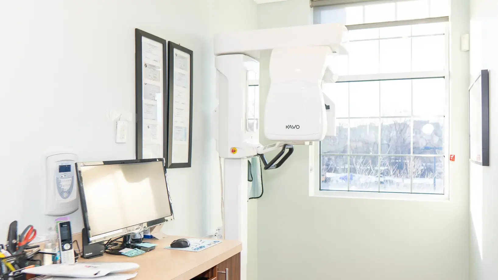 Dental X-ray machine in a brightly lit room with a computer and certificates on the wall.