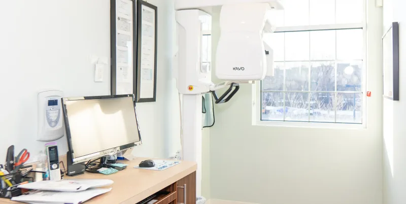 A dental X-ray machine is positioned next to a desk with a monitor.