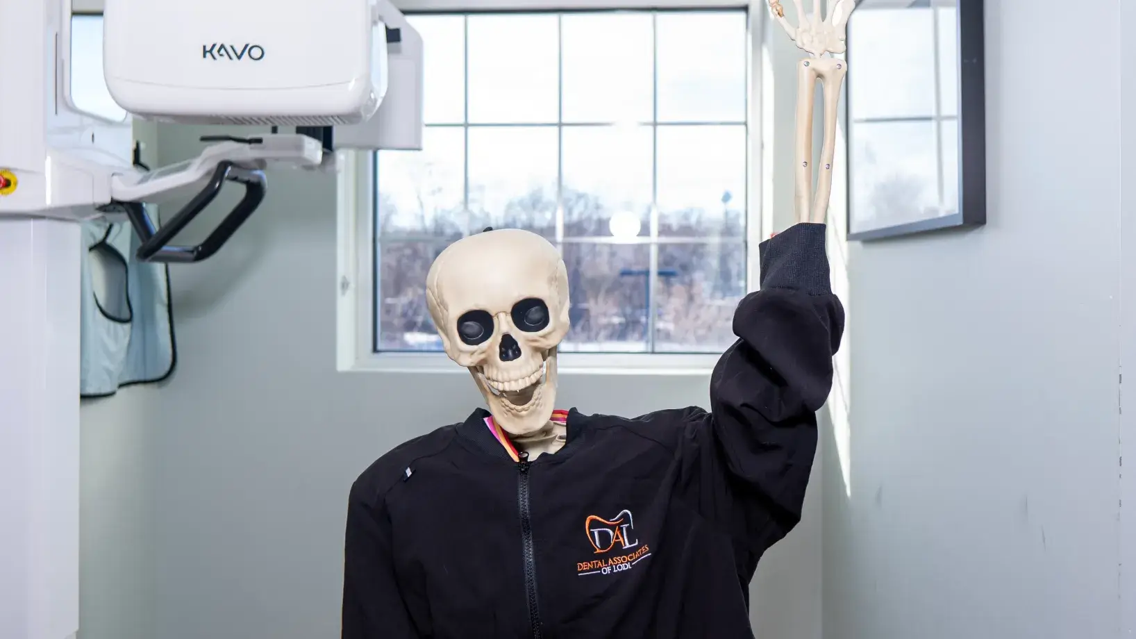 A skeleton wearing a black jacket is sitting on a chair, raising one arm.