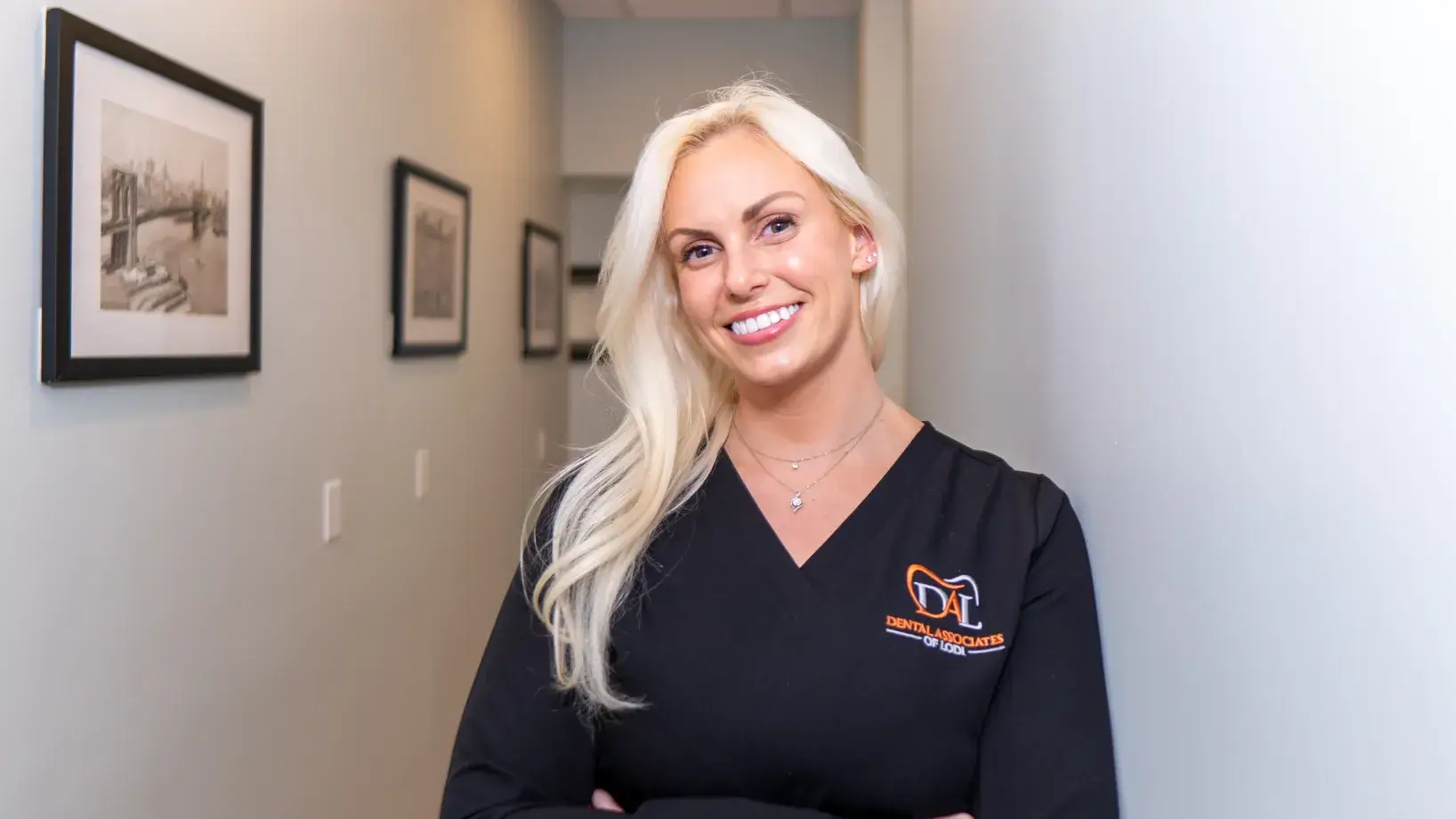 A smiling woman with long blonde hair stands in a hallway wearing a black uniform top.