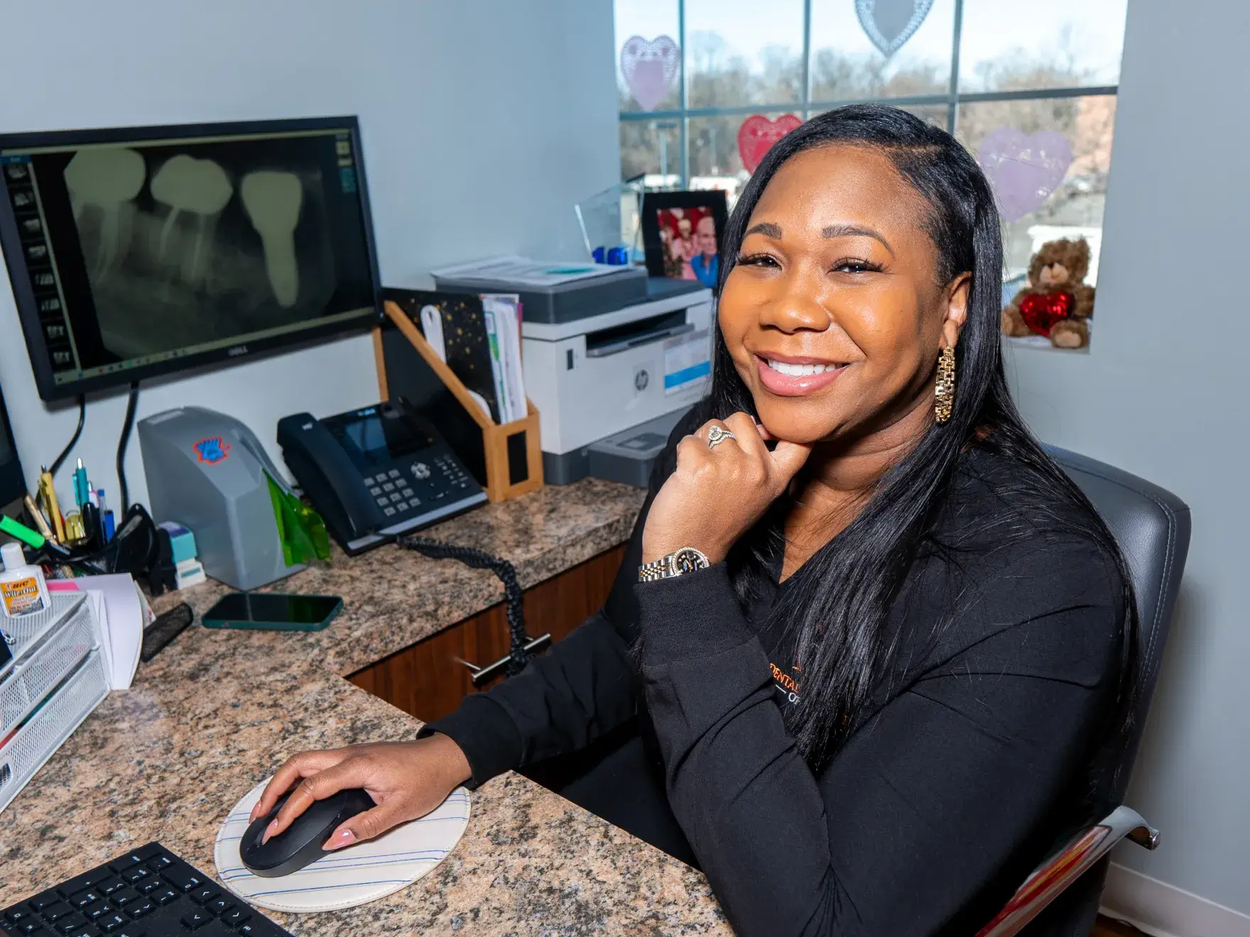 Smiling woman seated at a desk in an office with dental X-rays on a monitor behind her.